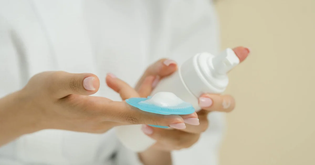 Hands hold a blue silicone gel sheet while another hand dispenses white gel from a bottle, demonstrating a non-silicone approach to post-tattoo scar care.