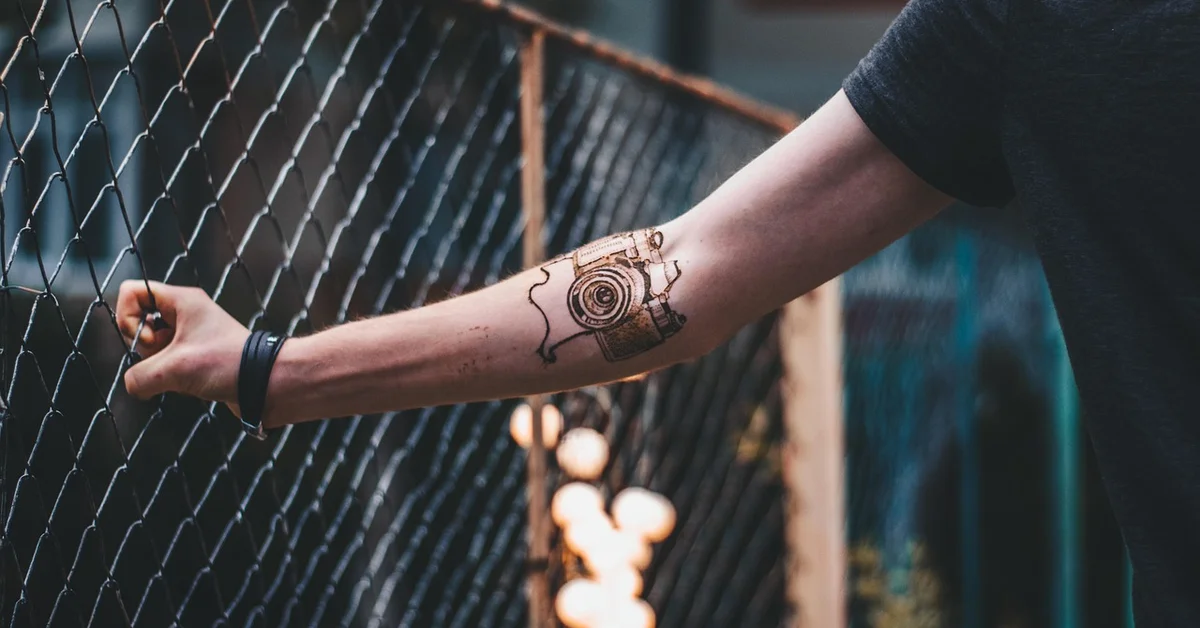 Close-up of a forearm tattoo as the arm grips a chain-link fence, with blurred lights in the background.