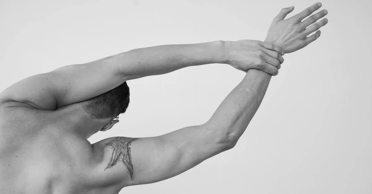 Black-and-white photo of a shirtless person with an upper-arm tattoo, arms raised and stretched overhead, focusing on placement and visibility