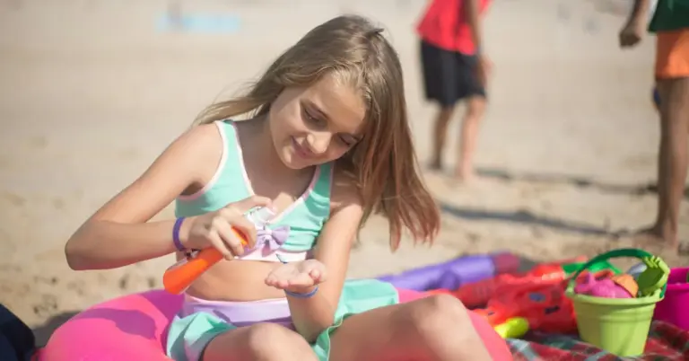 A young girl sits on a colorful beach blanket, applying sunscreen to her arm with an orange bottle, while beach toys and sand are in the background.