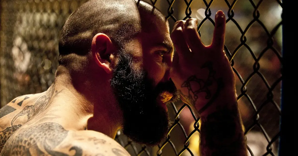 Close-up profile of a tattooed man with a full beard pressing his head against a chain-link fence, suggesting reflection and change after a breakup.
