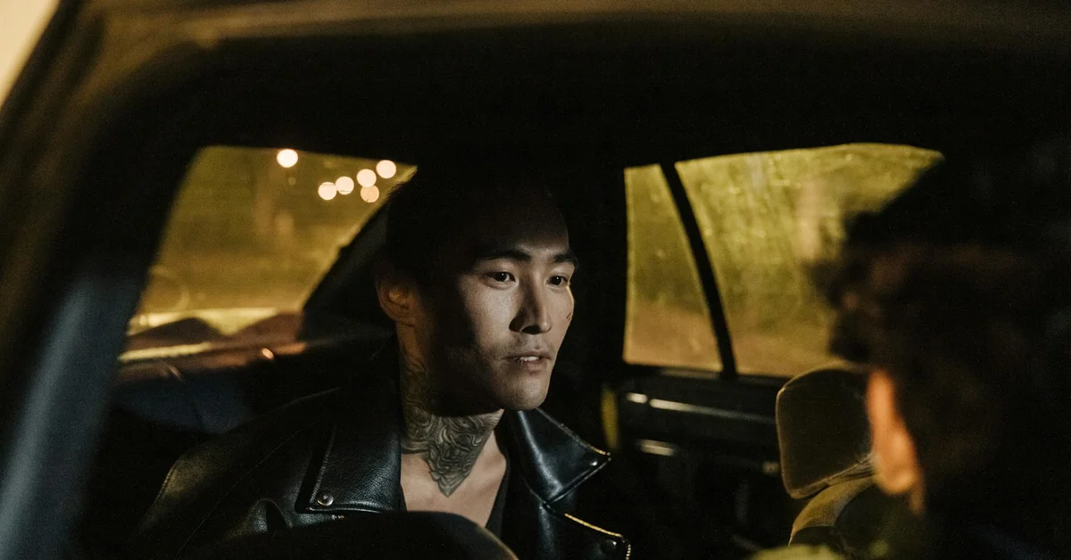 A man in a black leather jacket sits in the driver's seat of a car at night, with a blurred passenger nearby.
