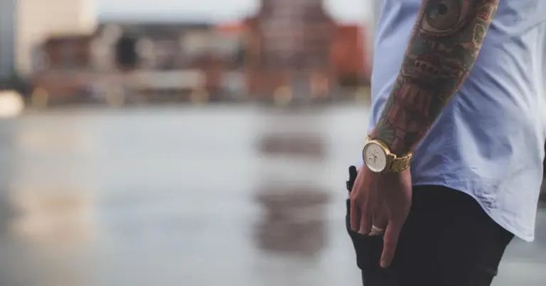 Close-up of a tattooed forearm resting by the side, wearing a light blue shirt sleeve and a gold watch.