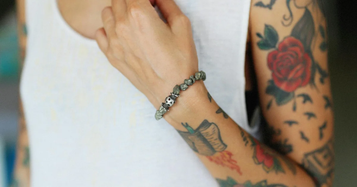 Close-up of a tattooed forearm with a red rose and other designs visible against a white sleeveless shirt