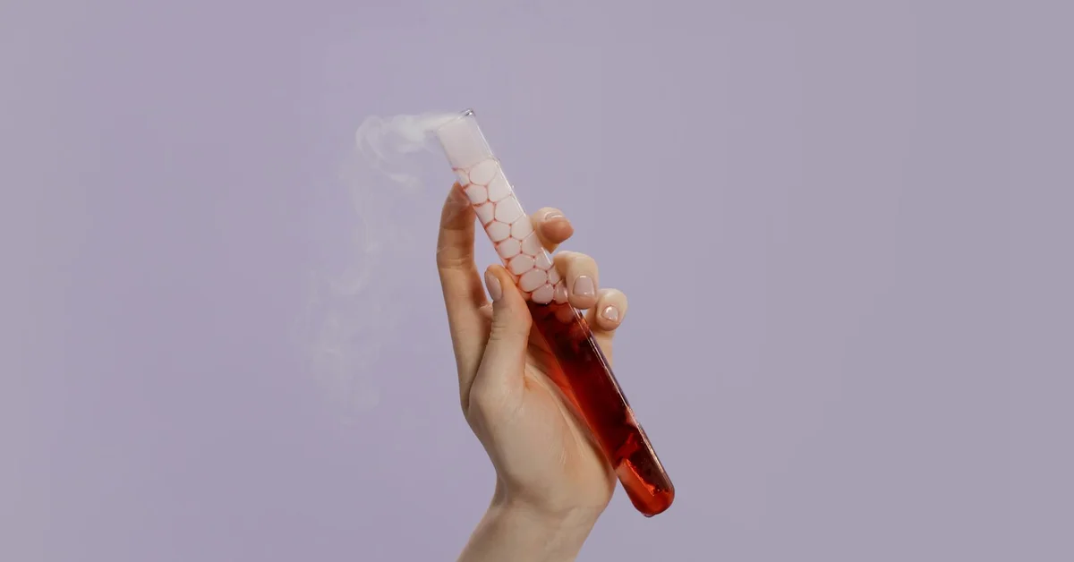 A hand holding a test tube with a red liquid and white crystalline material, emitting vapor, against a soft purple background.