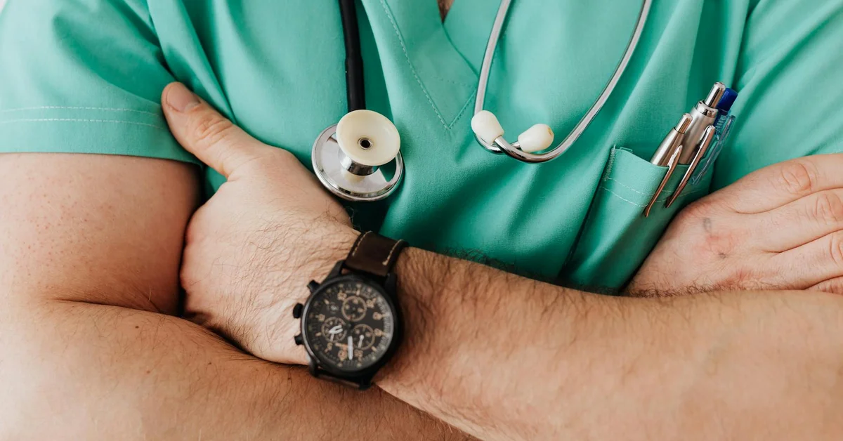 Medical professional in teal scrubs with a stethoscope and pens, arms crossed.