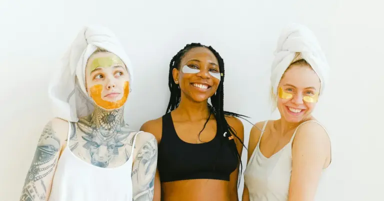 Three women with facial masks and towels wrapped around their heads, smiling in a bright studio.