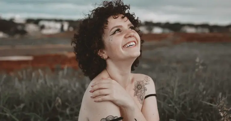 Young woman with curly hair outdoors, smiling, with a tattoo on her shoulder.