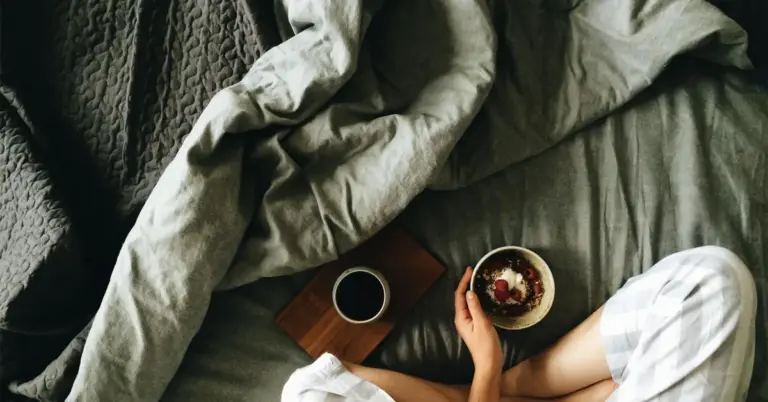 Top-down view of a person resting on a bed with a blanket, a cup of coffee on a wooden tray, and a bowl nearby.