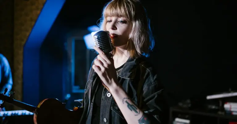 Female musician performing on stage, singing into a vintage microphone with a tattoo visible on her forearm.