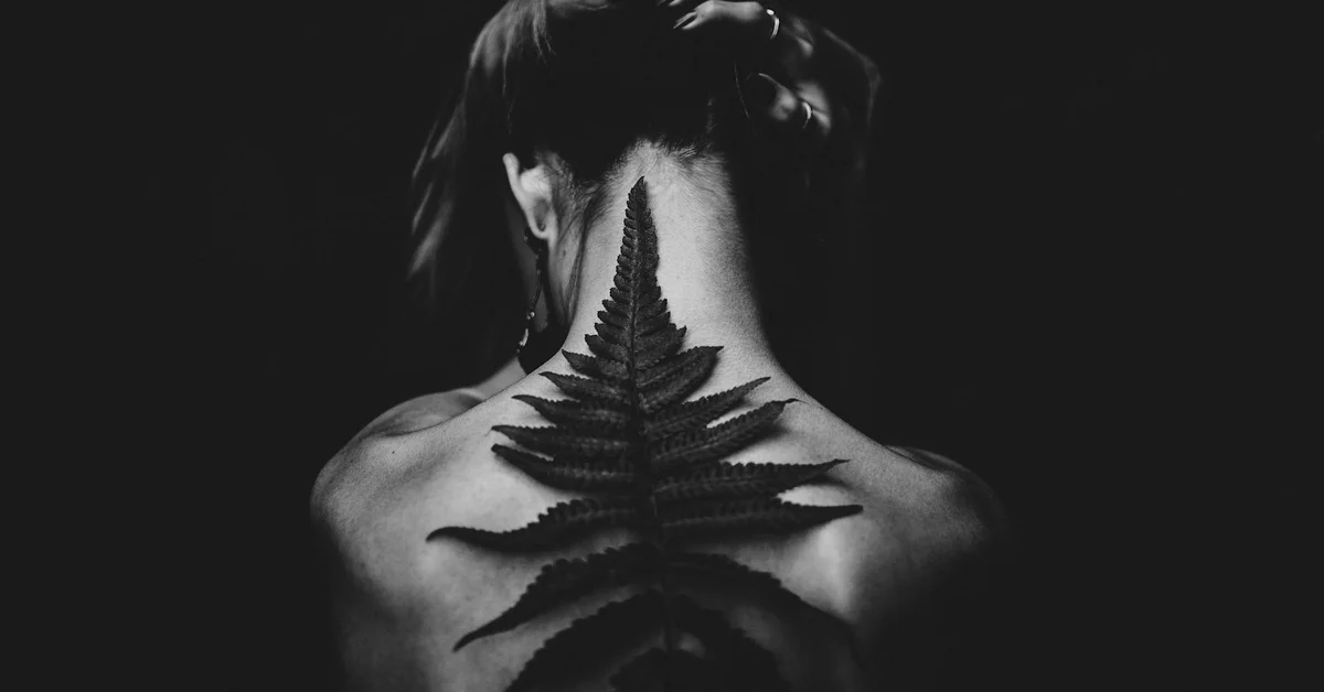 Back view of a person with a vertical fern-shaped tattoo along the spine, shown in black and white.