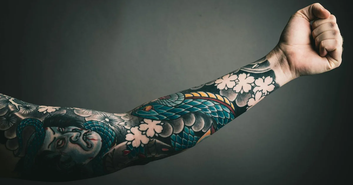 Close-up of a heavily tattooed forearm with a colorful traditional-style sleeve against a gray background
