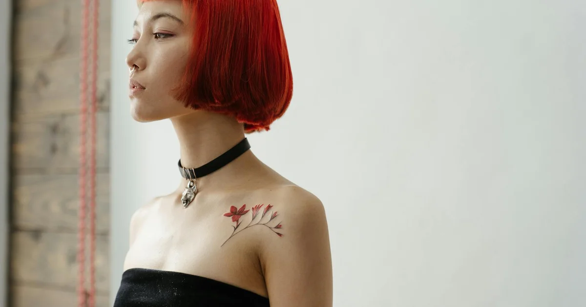 Profile of a person with short red hair and a small floral tattoo on the shoulder, wearing a black strapless top and a choker.