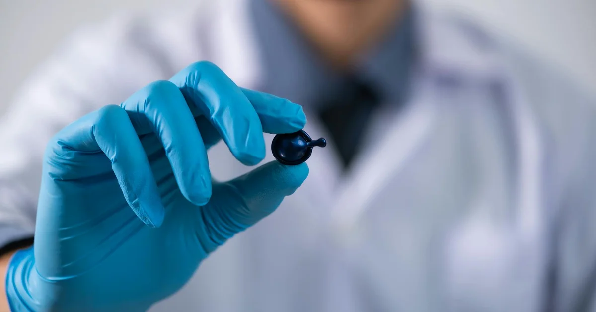 Close-up of a clinician's gloved hand holding a small bottle with a dropper in a clinical setting.