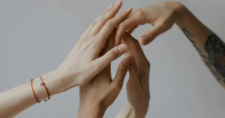 Close-up of two people’s hands; one wrist shows a tattoo and both wear bracelets, against a neutral background.