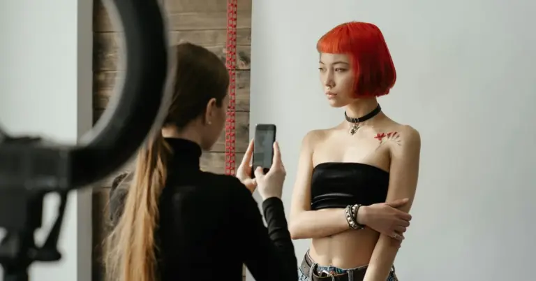 Portrait session in a studio: a model with a bright red bob hairstyle posed while a photographer takes a photo, with a measuring tape visible in the background.