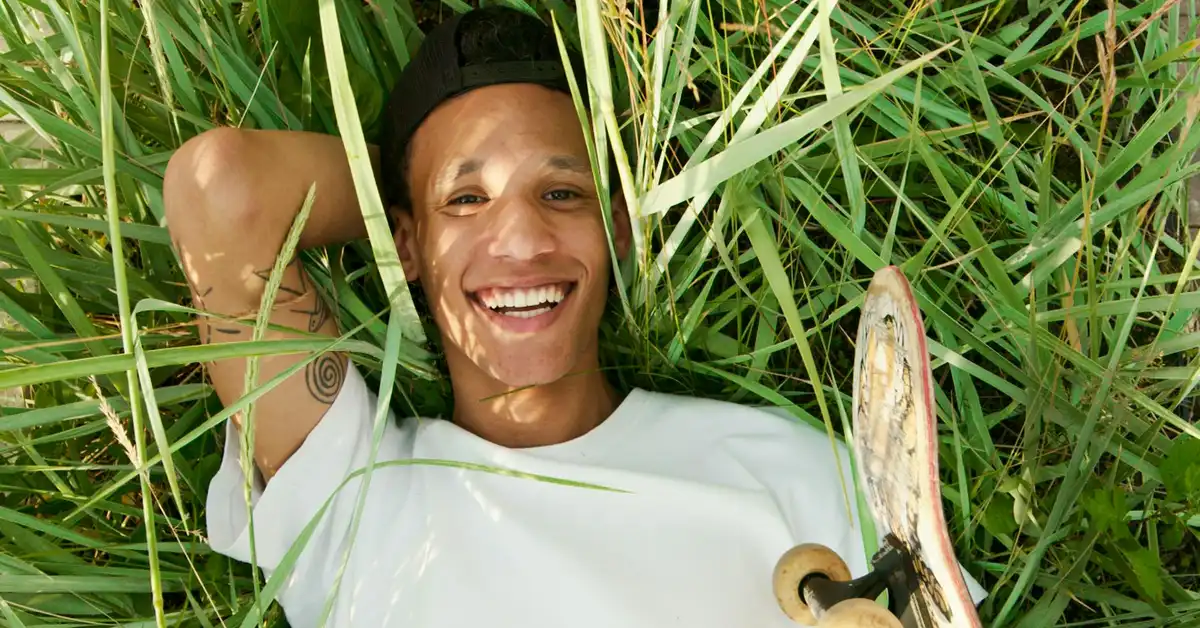 Person lying in tall grass outdoors with a skateboard nearby, smiling at the camera