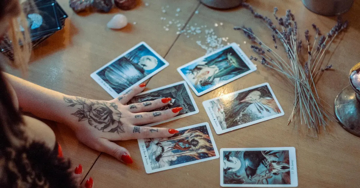 A tattooed hand with red-painted nails reaches toward several illustrated cards spread on a wooden table, with dried lavender and other objects in the background.