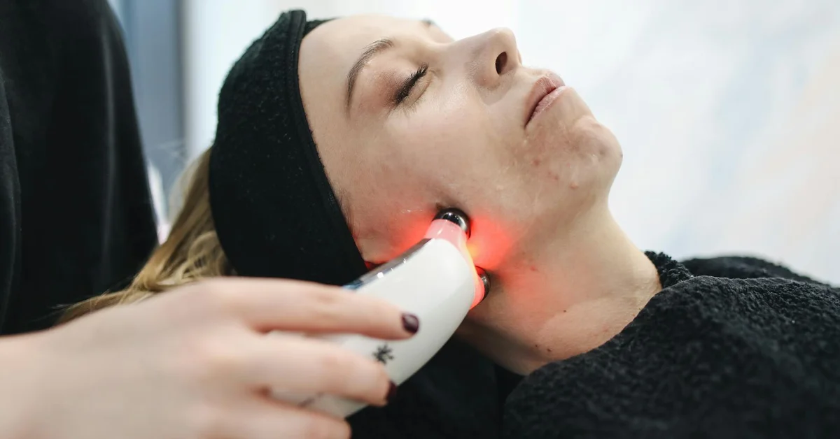 A clinician applies a handheld laser device to a patient’s neck during a cosmetic treatment; a red light emanates from the device as the patient rests with a black headband.