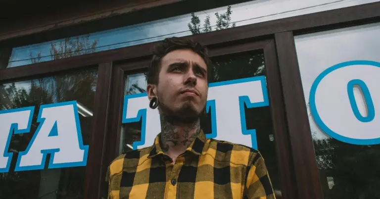 A tattooed man wearing a yellow and black plaid shirt stands in front of a storefront with large blue lettering in the window.