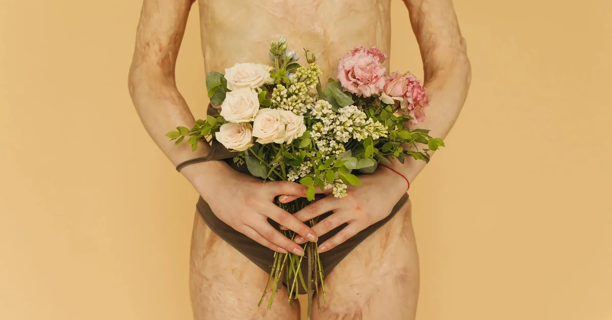 A person wearing black underwear stands against a peach background, arms around a bouquet of flowers covering the midsection, showing visible stretch marks and scarring on the torso.