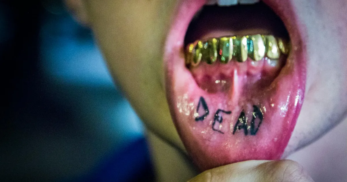 Close-up of an open mouth showing a tongue tattoo that reads 'DEAN' with gold grill teeth.