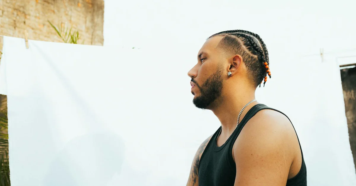Profile view of a man with cornrow hair wearing a black sleeveless shirt outdoors against a light wall