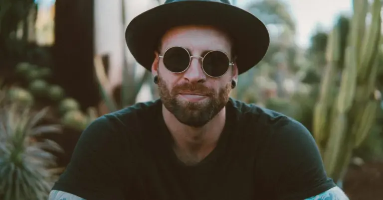 A man wearing a black shirt, round sunglasses, and a wide-brimmed hat stands outdoors, with plants in the background.