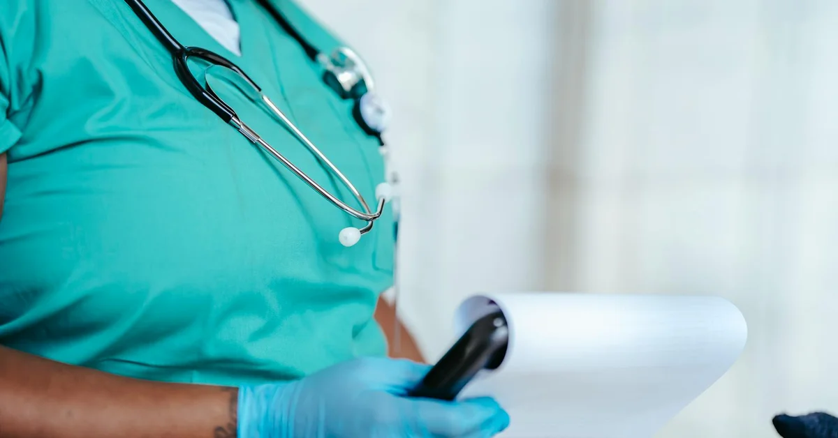 Medical professional wearing teal scrubs and gloves holding a clipboard with notes and a stethoscope around their neck.