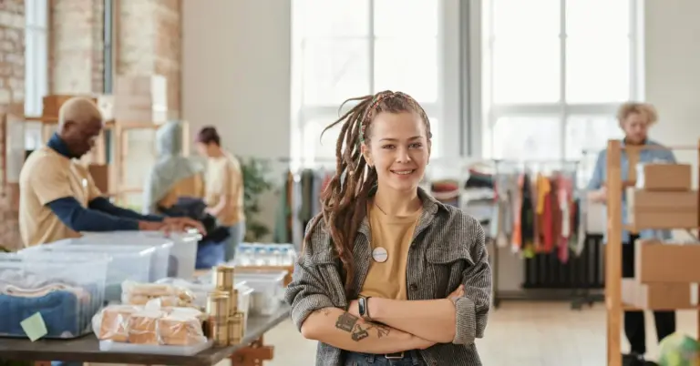 Smiling woman with tattoos on her forearm stands with arms crossed in a bright, open studio. Other people and racks of clothing are visible in the background.