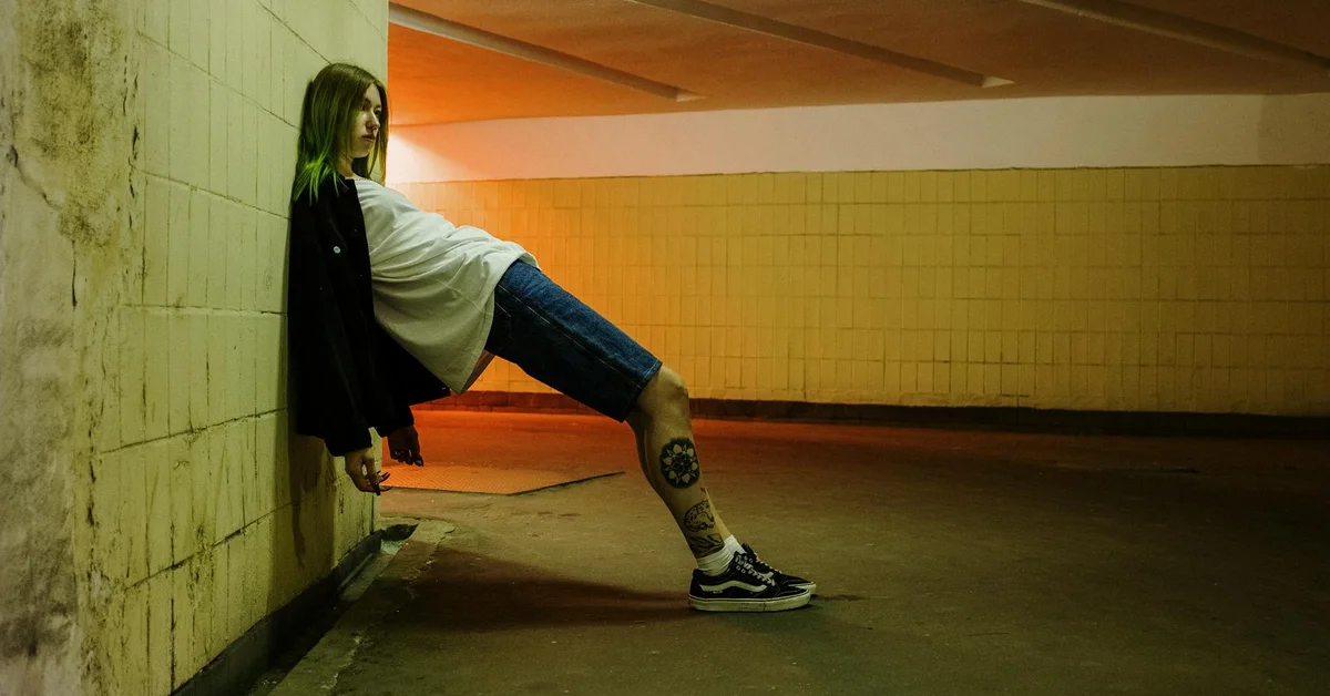 A person with a leg tattoo leans against a wall in a dimly lit tunnel.