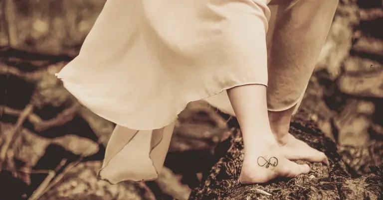 Barefoot ankle with an infinity tattoo, draped by a flowing dress, standing on a rocky surface.