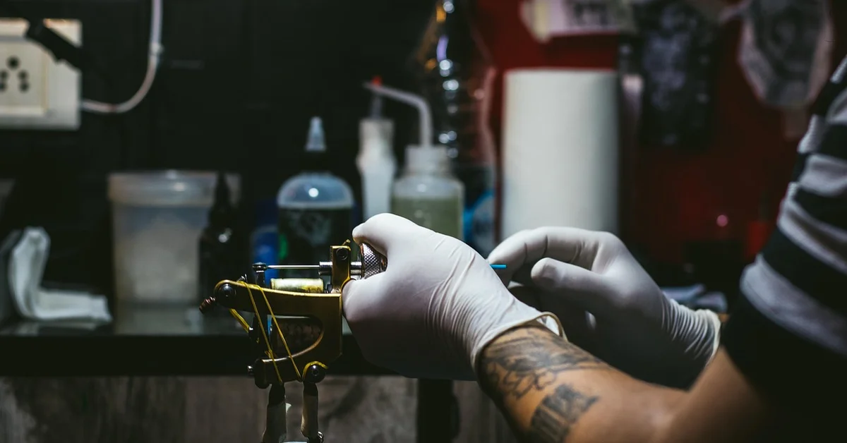 Close-up of a tattoo removal device being operated by gloved hands in a clinical setting, with equipment and supplies visible in the background.