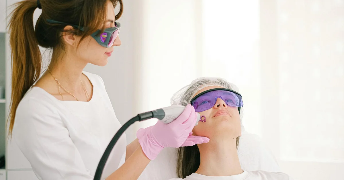 Clinician in white coat and pink gloves performing a pico laser treatment on a patient who wears protective eyewear; both are in a clinical setting with a handheld laser device.