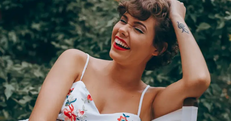 Smiling woman with short hair wearing a white floral dress outdoors, with a small tattoo on her forearm
