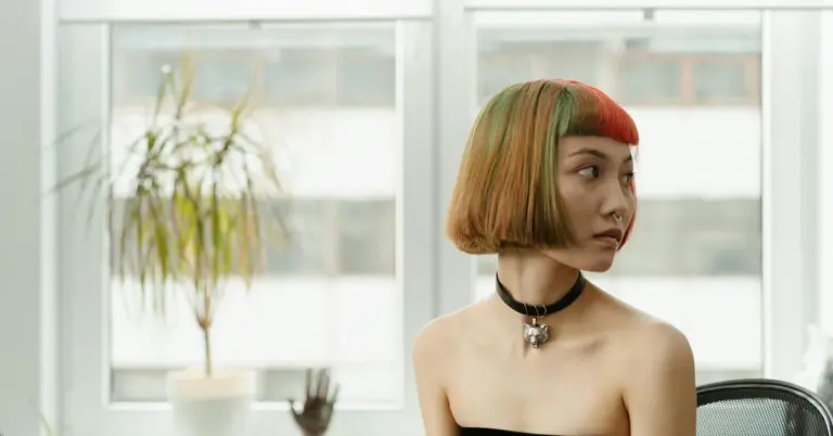 Portrait of a person with a short, multicolored bob haircut wearing a black choker, seated indoors by a window.