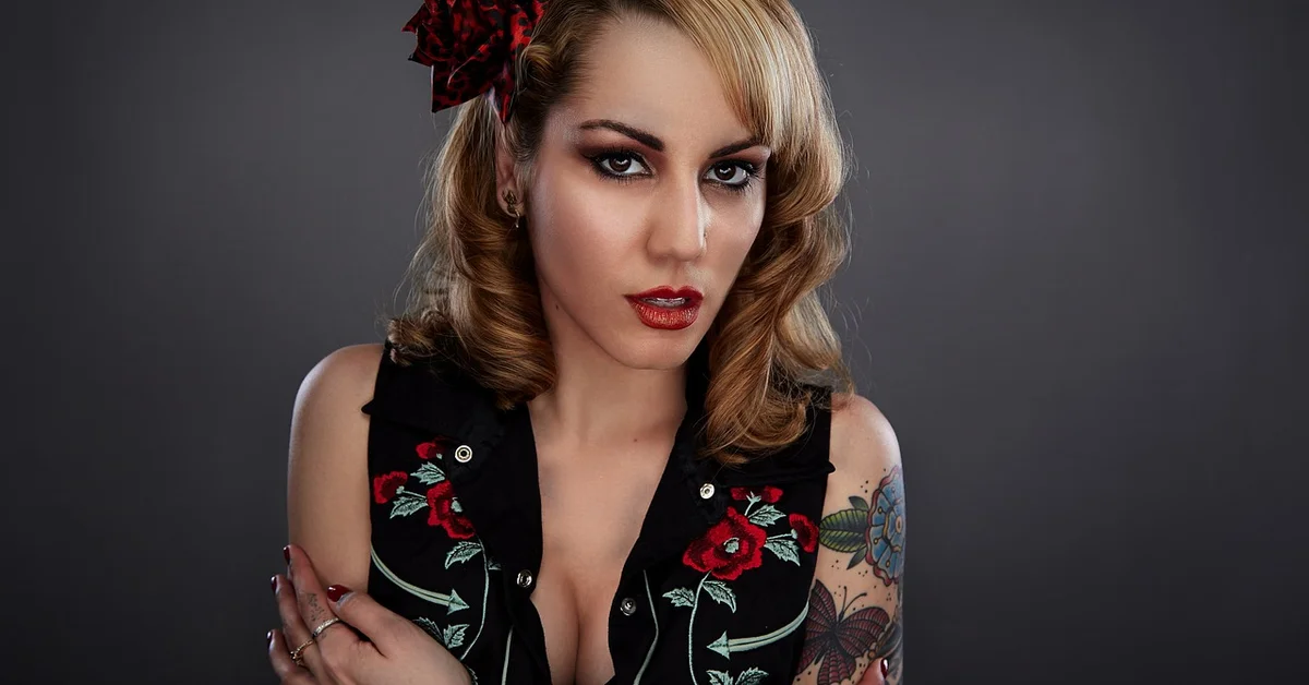 Portrait of a woman with blonde hair, red lipstick, and visible tattoos on her left arm, wearing a black floral vest, posed against a gray background.