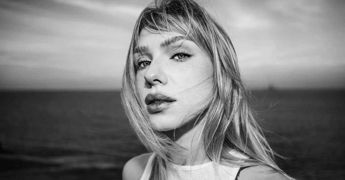Black-and-white portrait of a woman by the sea with windswept hair, looking at the camera