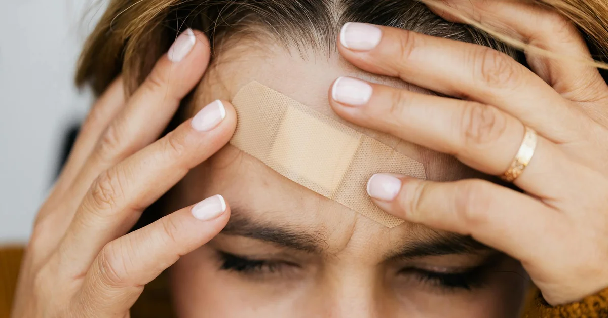 Close-up of a person with a bandage on their forehead, hands near the bandage