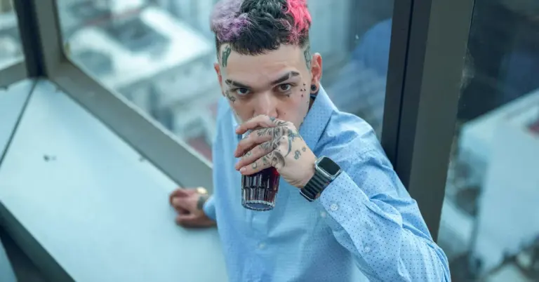 Young person with colorful hair and tattoos, wearing a light blue shirt, leaning on a windowsill and drinking from a can