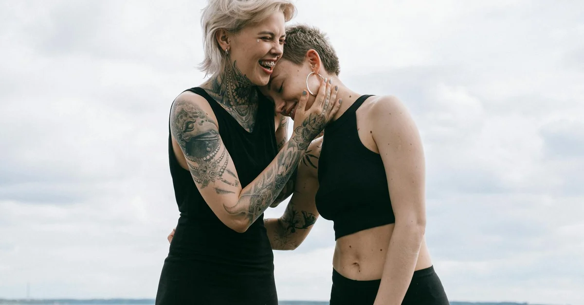 Two tattooed women in black athletic wear share a moment outdoors, smiling and touching faces.