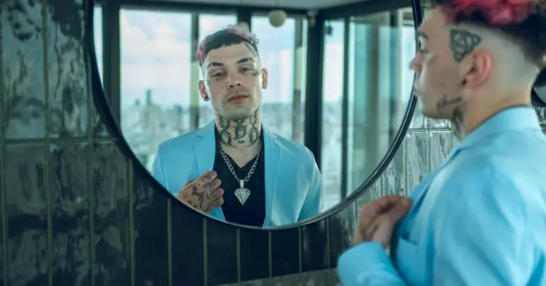 Tattooed person wearing a light blue blazer looks at their reflection in a round mirror in a modern bathroom.