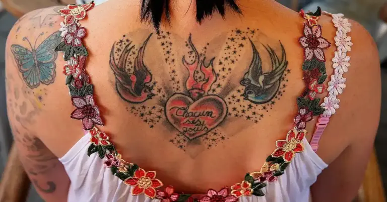 Back view of a person with multiple tattoos: a central heart with two birds on either side, various ink designs on the shoulders, and a floral garment along the upper back.
