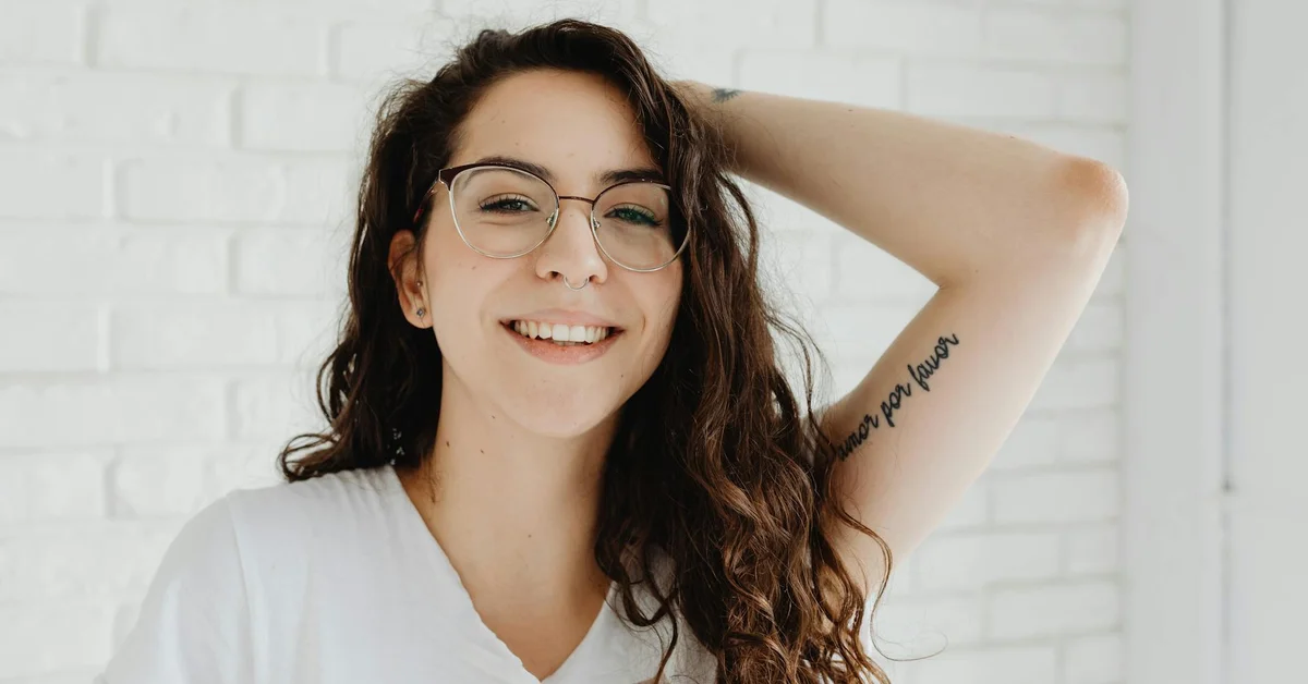 Young woman with glasses smiling, showing a forearm tattoo, standing against a white brick wall indoors.