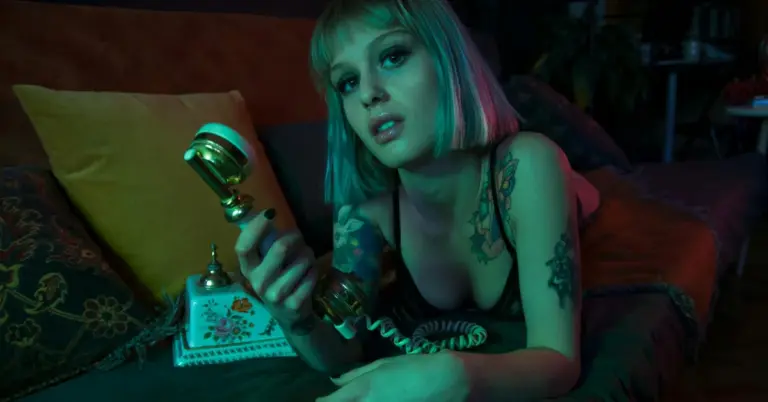 Tattooed person lying on a couch under teal lighting, holding a vintage rotary phone.