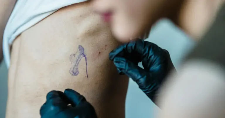 Close-up of gloved hands preparing to tattoo on the upper arm where stretch marks are visible, with a purple outline drawn on the skin.