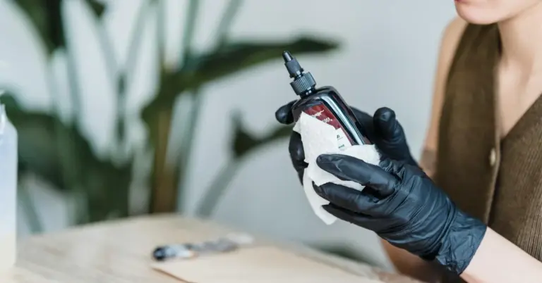 A gloved hand holding a bottle of tattoo ink with a dropper cap on a light wooden surface.