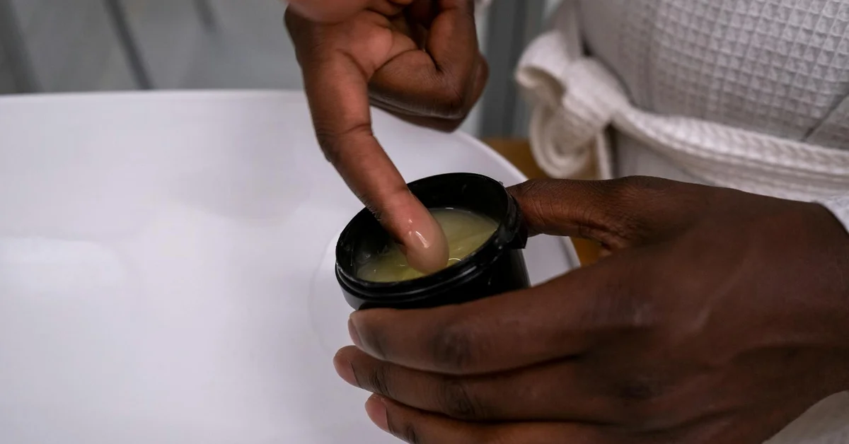 Close-up of hands applying a small amount of ointment from an open jar onto the skin