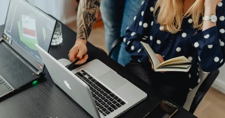 Two people with visible tattoos sit at a desk with laptops and a notebook, planning a tattoo removal business.