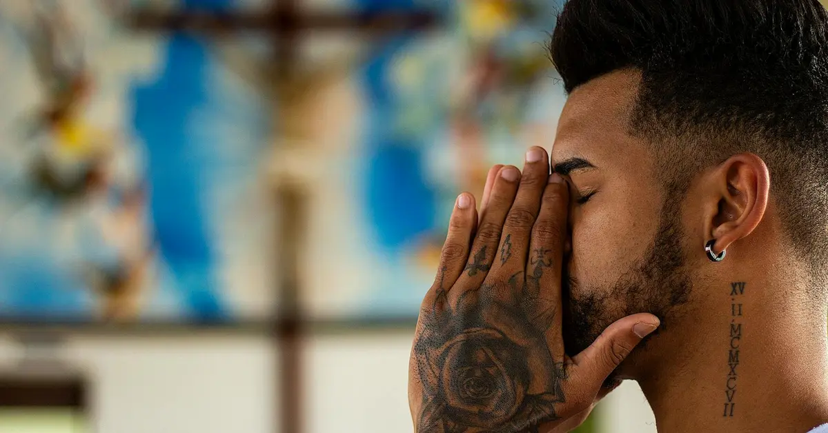 Close-up side profile of a tattooed man with his hands over his face, visible tattoos on his hand and neck.